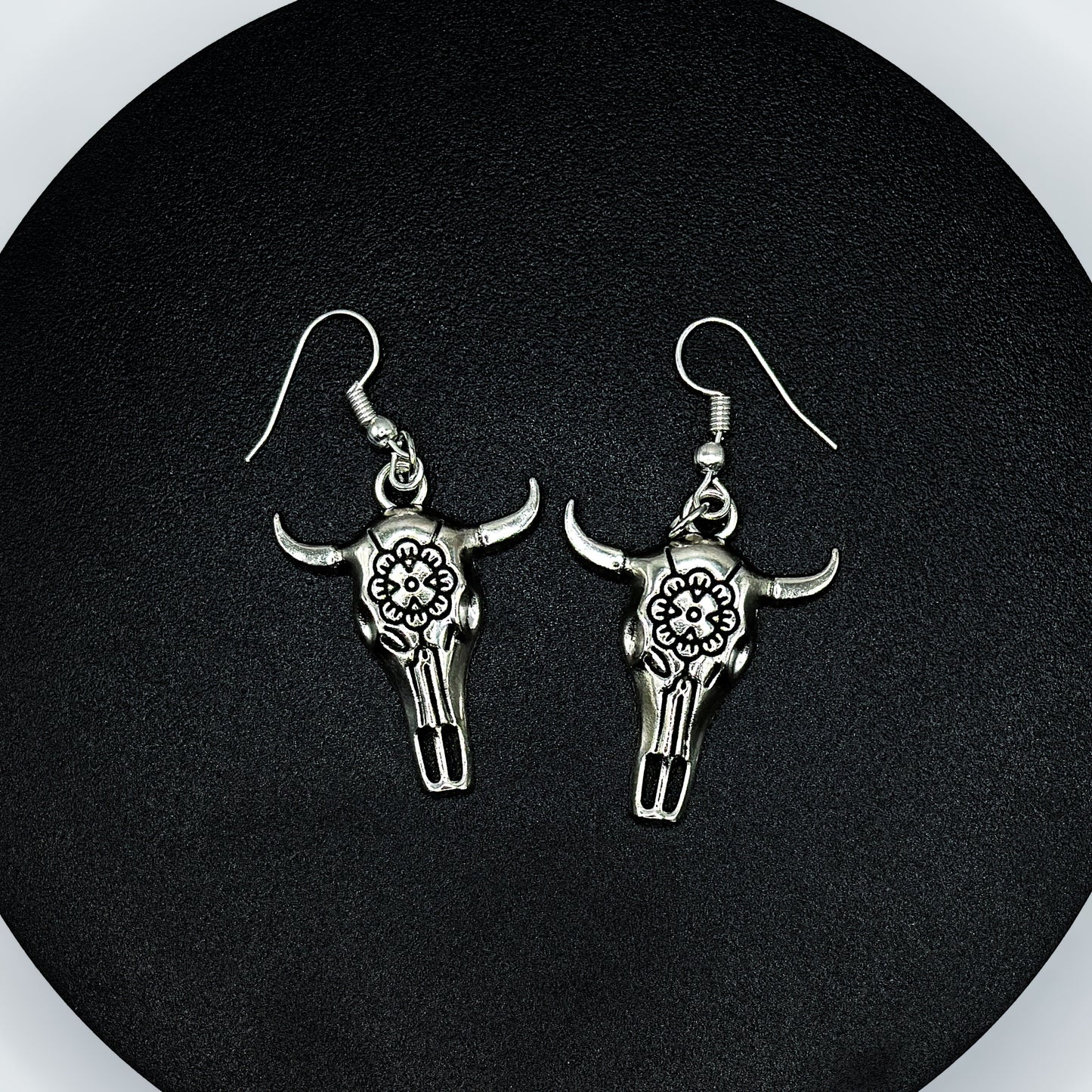 Pair of antique silver bull skull drop earrings with floral engraving on a black display stand