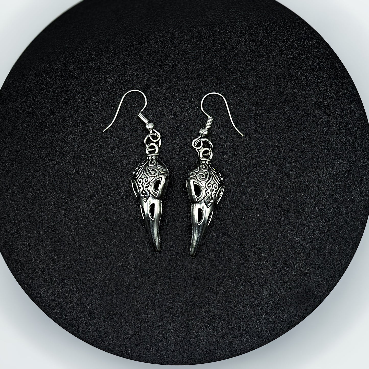 Pair of antique silver raven skull drop earrings with ornate scrollwork on a black display stand
