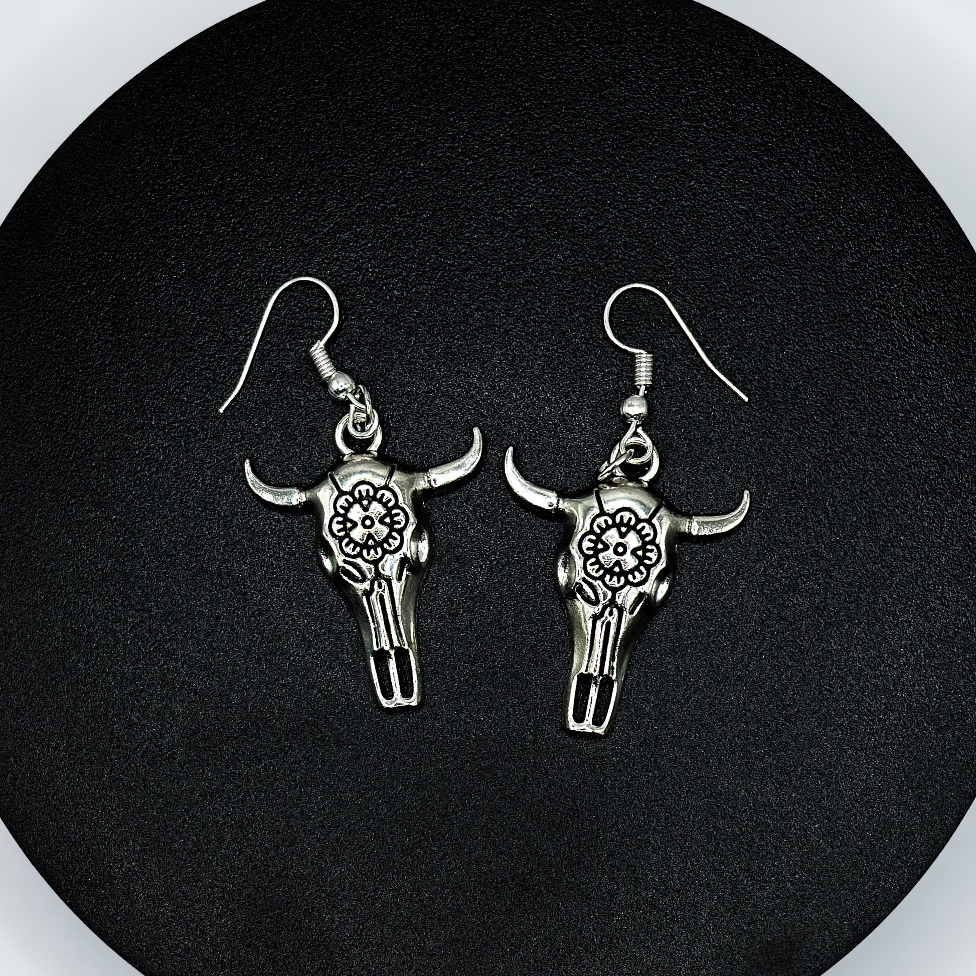Pair of antique silver bull skull drop earrings with floral engraving on a black display stand